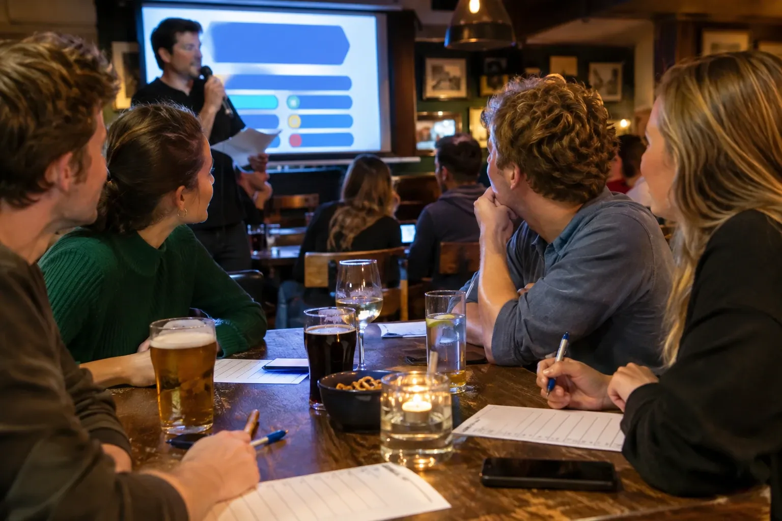 Groep spelers in pub rond tafel met quizformulieren, drankjes en presentator op scherm